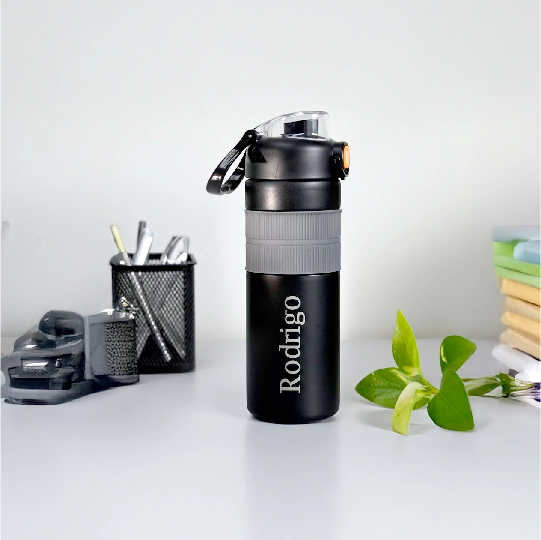 Garrafa térmica preta personalizada com o nome Rodrigo gravado a laser, apoiada sobre uma mesa de trabalho ao lado de um porta-canetas. Exemplo de brinde corporativo útil na rotina de escritório.